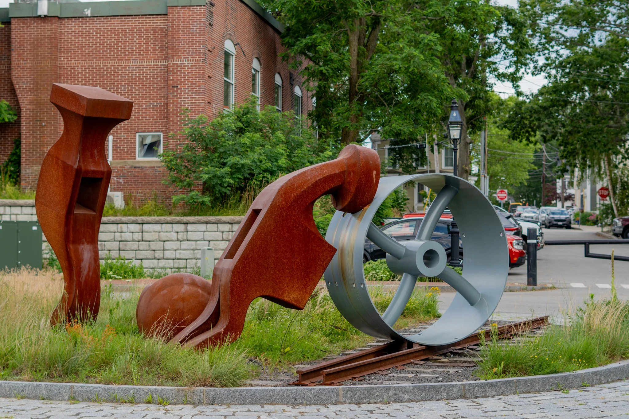 Outdoor sculpture consisting of industrial pieces, including a rust colored hammer head, a metal wheel, and a ball, situated on a small grassy area near a sidewalk with a brick building, trees, and parked cars in the background.