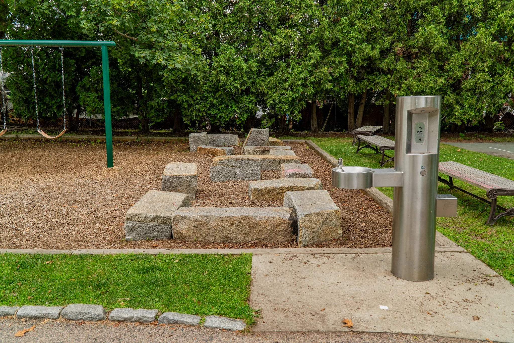 A public park features a playground area with a swing set on the left, a curved stone seating arrangement in the center, a drinking fountain on the right, and benches along the background, all surrounded by green trees and grass.