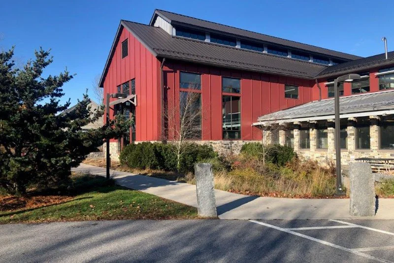 Red and gray modern building with sloped roof, surrounded by shrubbery and trees, with a clear blue sky.