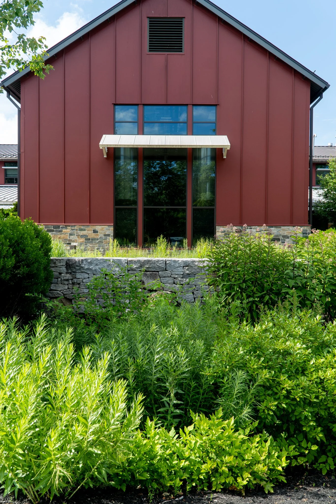 Front view of a modern building with a red exterior, stone accents at the base, large glass windows, and surrounded by lush green plants and shrubs.