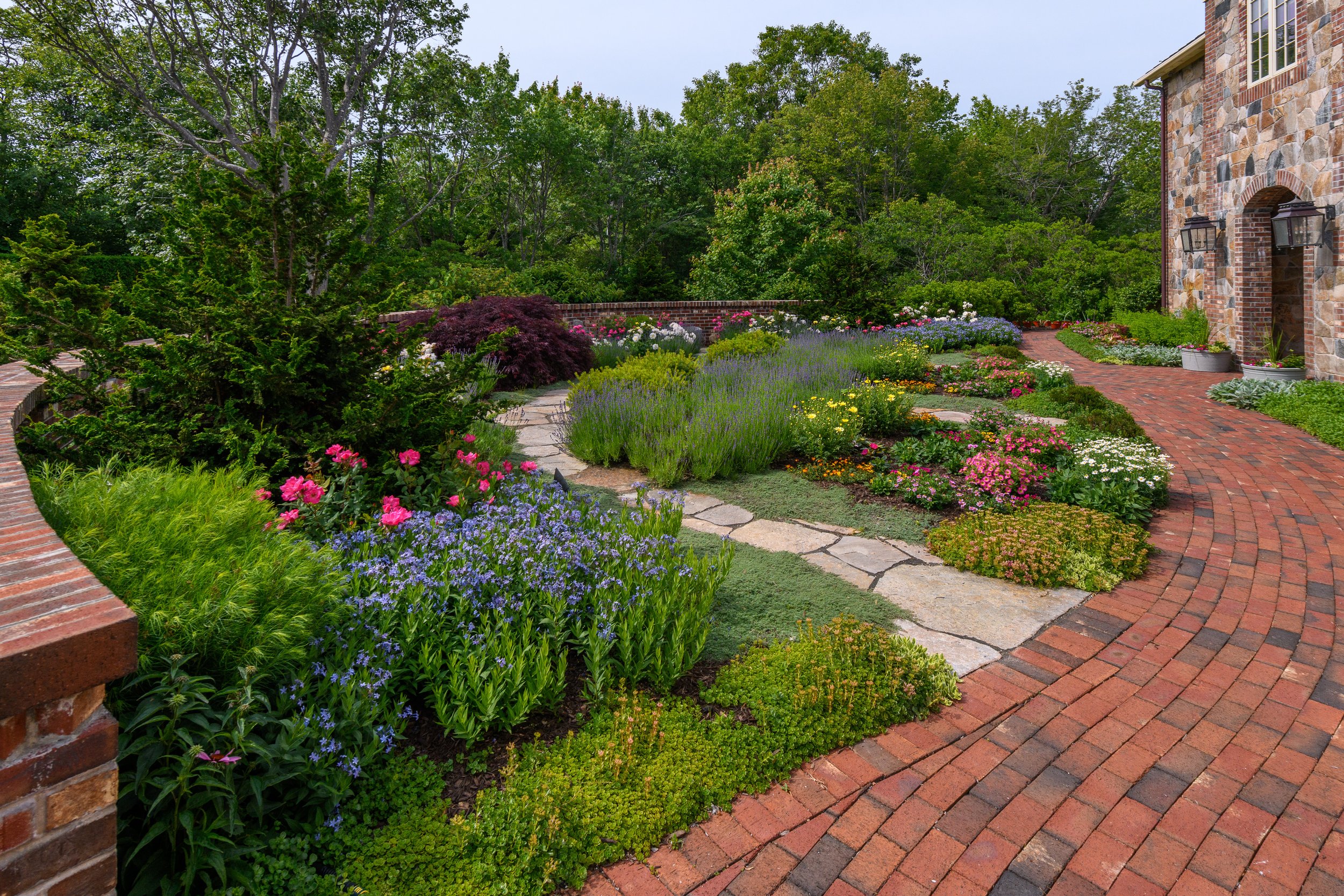 A lush garden with colorful flowers, a stone pathway, and a brick house wall in the background.