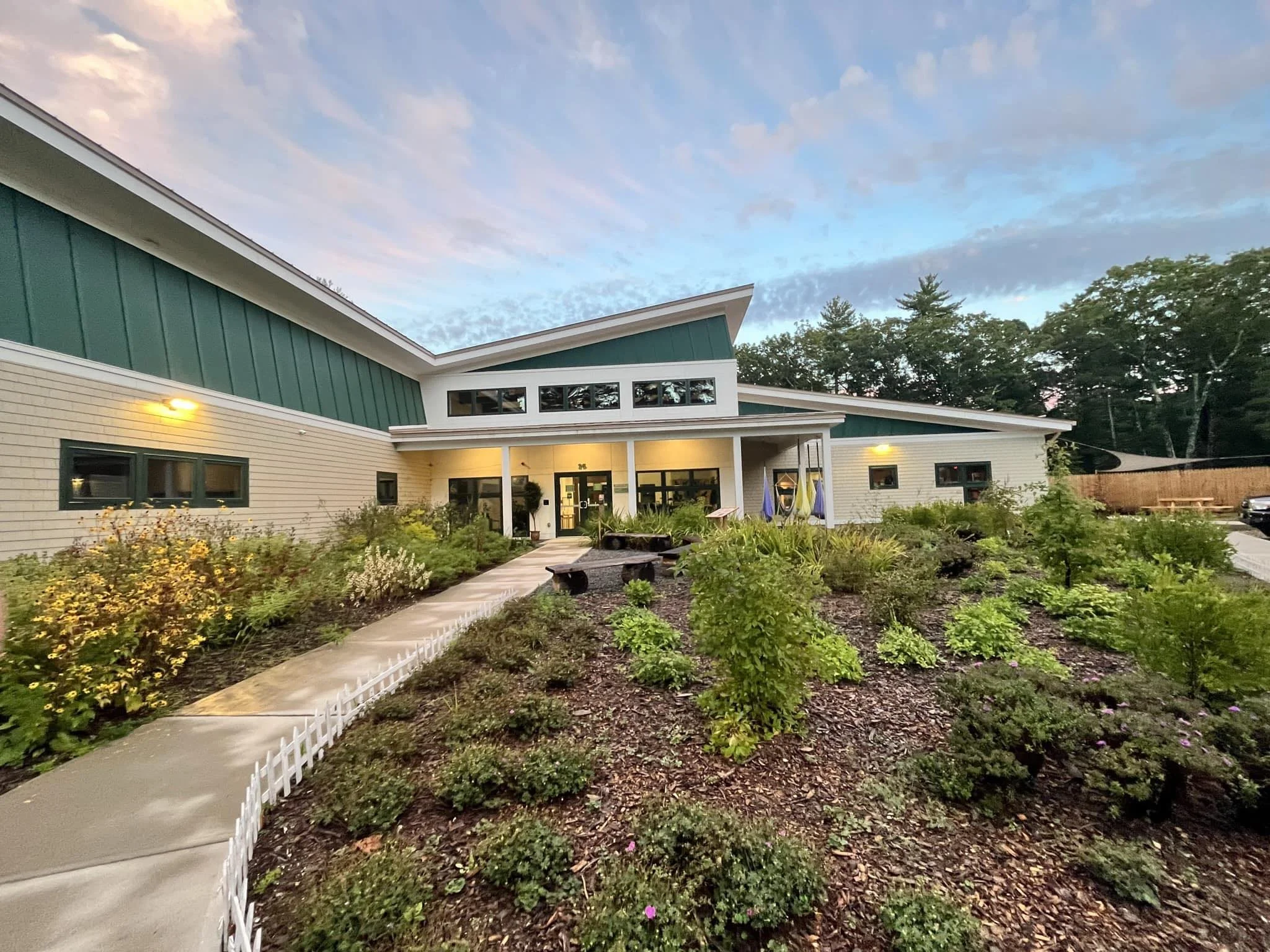 A modern building with a lush landscaped front yard and a concrete pathway leading to the entrance, illuminated by exterior lighting during dusk.