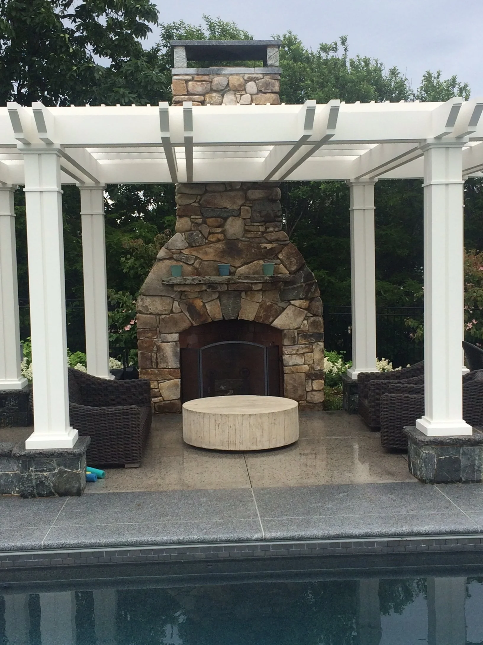 Outdoor seating area with a stone fireplace, white pergola, and wicker chairs next to a pool.