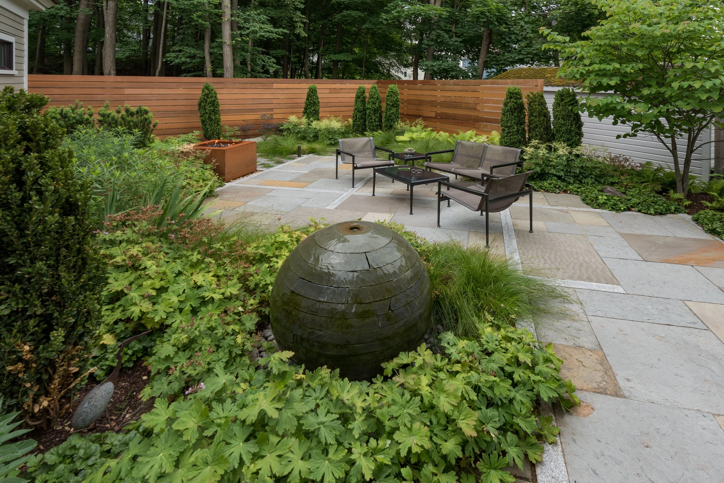 A backyard patio with stone paving, a water fountain, outdoor sofa and chairs, surrounded by green plants, trees, and a wooden privacy fence.