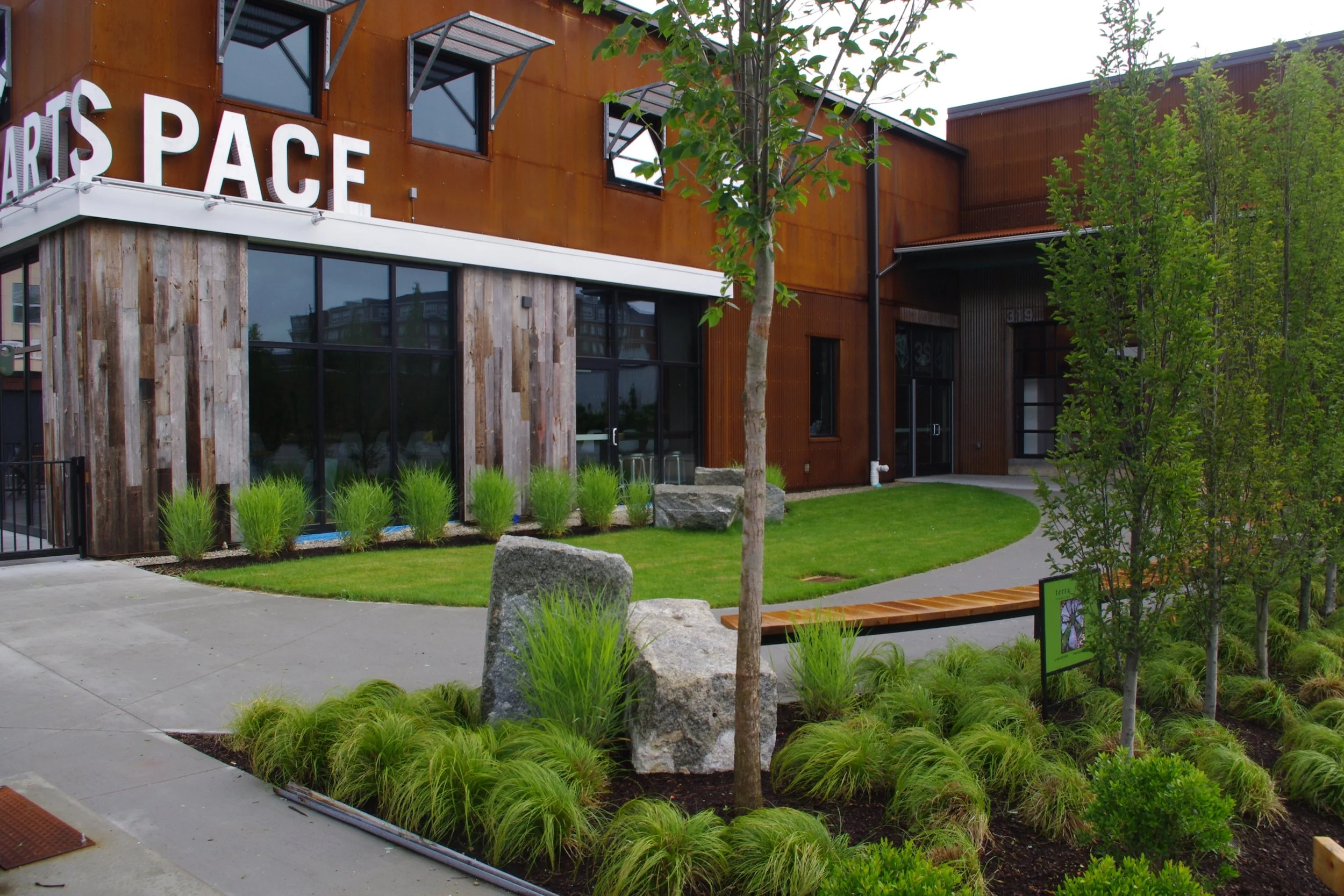 Exterior view of a modern arts space with a mix of wood and rust-colored metal siding, large windows, landscaped lawn with rocks, trees, shrubs, and a curved walkway.