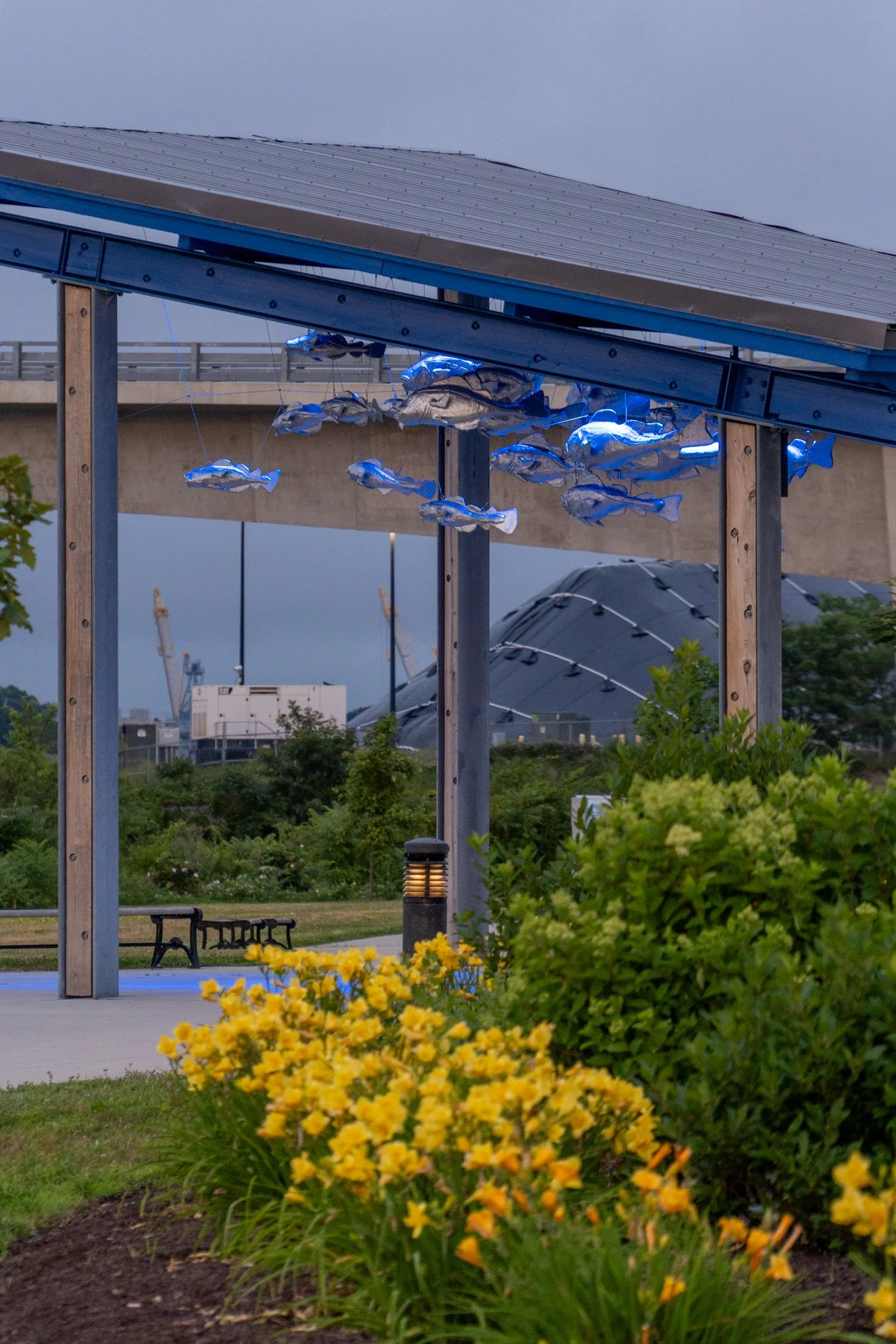 Outdoor scene with a metal and wood structure featuring hanging fish sculptures illuminated with blue lights, a park bench, greenery, yellow flowers, and industrial cranes in the background.