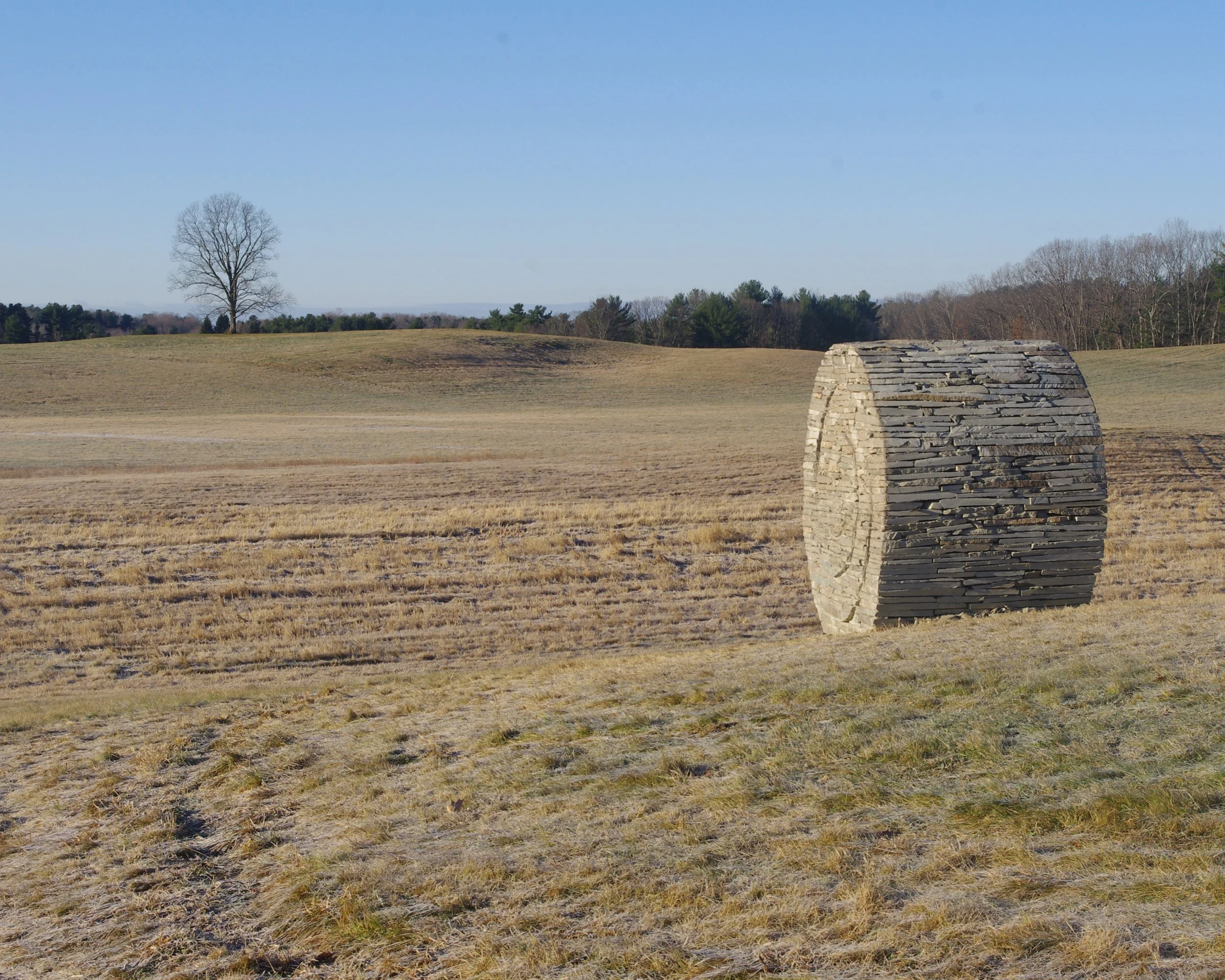 A large stone hay bale sculpture in a vast, open field with grassy terrain and a lone tree in the background under a clear, blue sky.