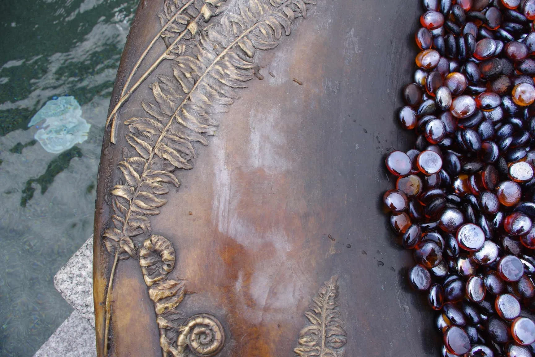 Close-up of a bronze fountain with decorative fern leaves relief, with a pile of dark purple fire stones on one side and a water surface reflecting in the background.
