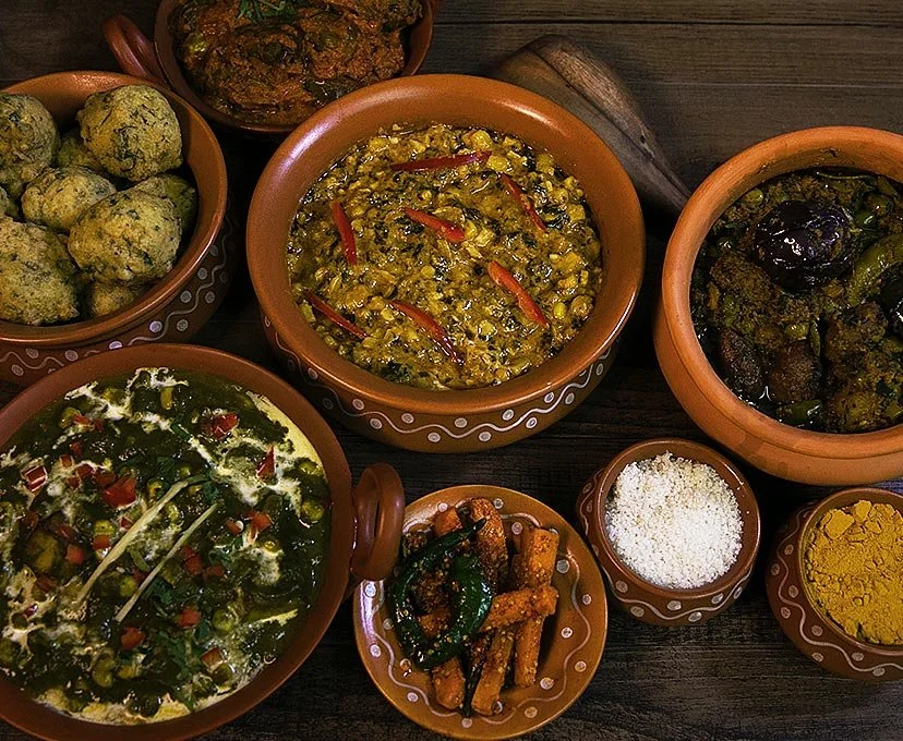 Assorted Indian dishes in clay pots, including vegetable fritters, a lentil curry, cooked eggplant, spinach Sabzi, dried green chilies, grated cheese, and turmeric powder.