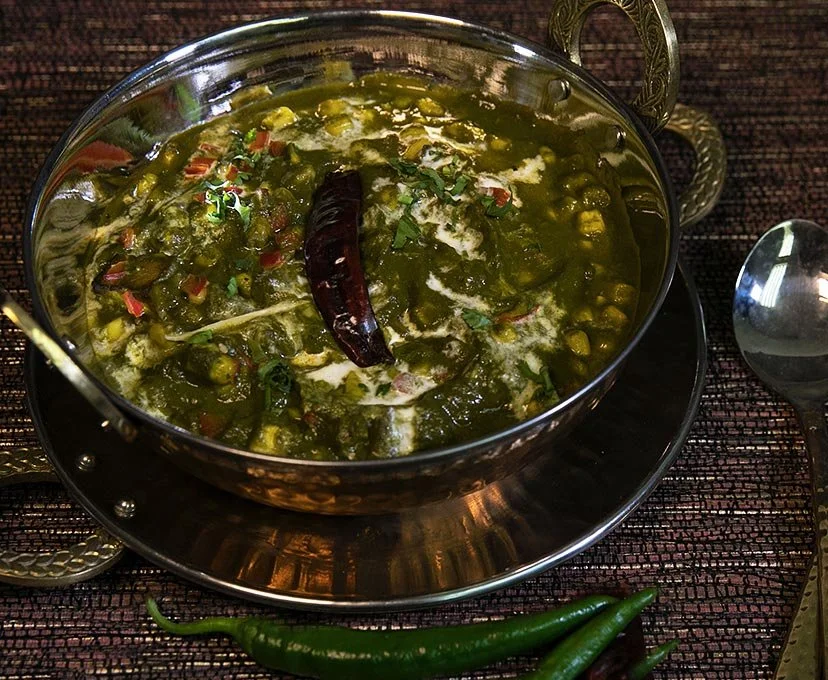 A bowl of Indian green curry with a dried red chili on top, garnished with cilantro, served on a metal tray with a green chili and a spoon nearby.