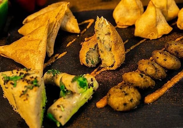 Assorted Indian samosas and paneer tikka on a black plate with green chutney and orange sauce