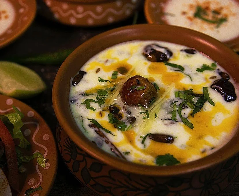 A close-up of Indian dish 'Dahi Bhalla' garnished with herbs, surrounded by bowls of similar dishes and lime wedges.