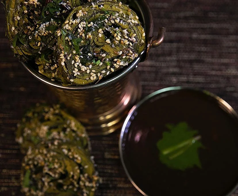 Close-up of two homemade green jalapeño pepper pickles coated with sesame seeds in a metal container and a small bowl of dark dipping sauce with a green leaf.