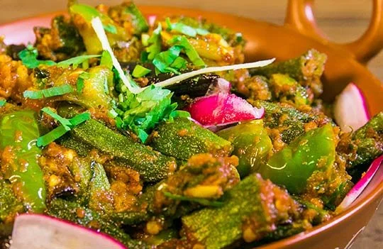 Close-up of a bowl of spicy okra stir-fry garnished with chopped herbs and radish slices.