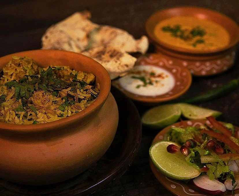 Close-up of traditional Indian meal with a bowl of seasoned rice, naan bread, small bowls of creamy soups or sauces, lime wedges, and a plate of salad with lime, pomegranate seeds, and vegetables on a dark wooden surface.
