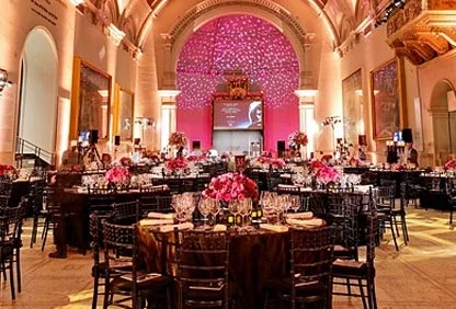 Elegant banquet hall decorated with pink floral centerpieces and large pink balloon arches, set for a formal event with tables, chairs, and uplighting.
