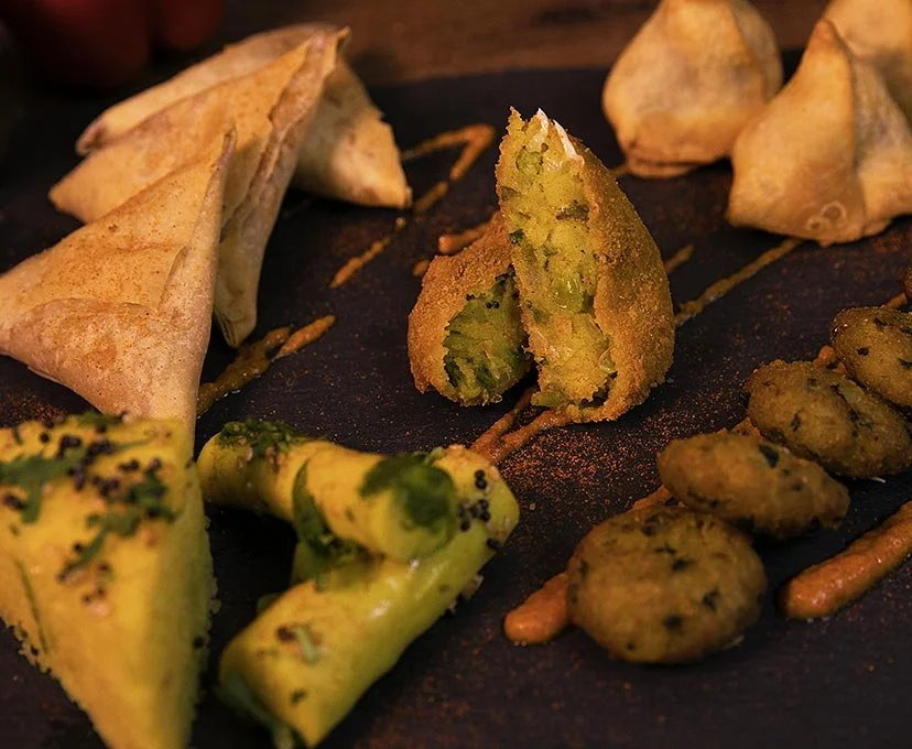 Assorted fried Indian snacks, including samosas, pakoras, and stuffed green chilies, garnished with spices and sauces on a black serving plate.