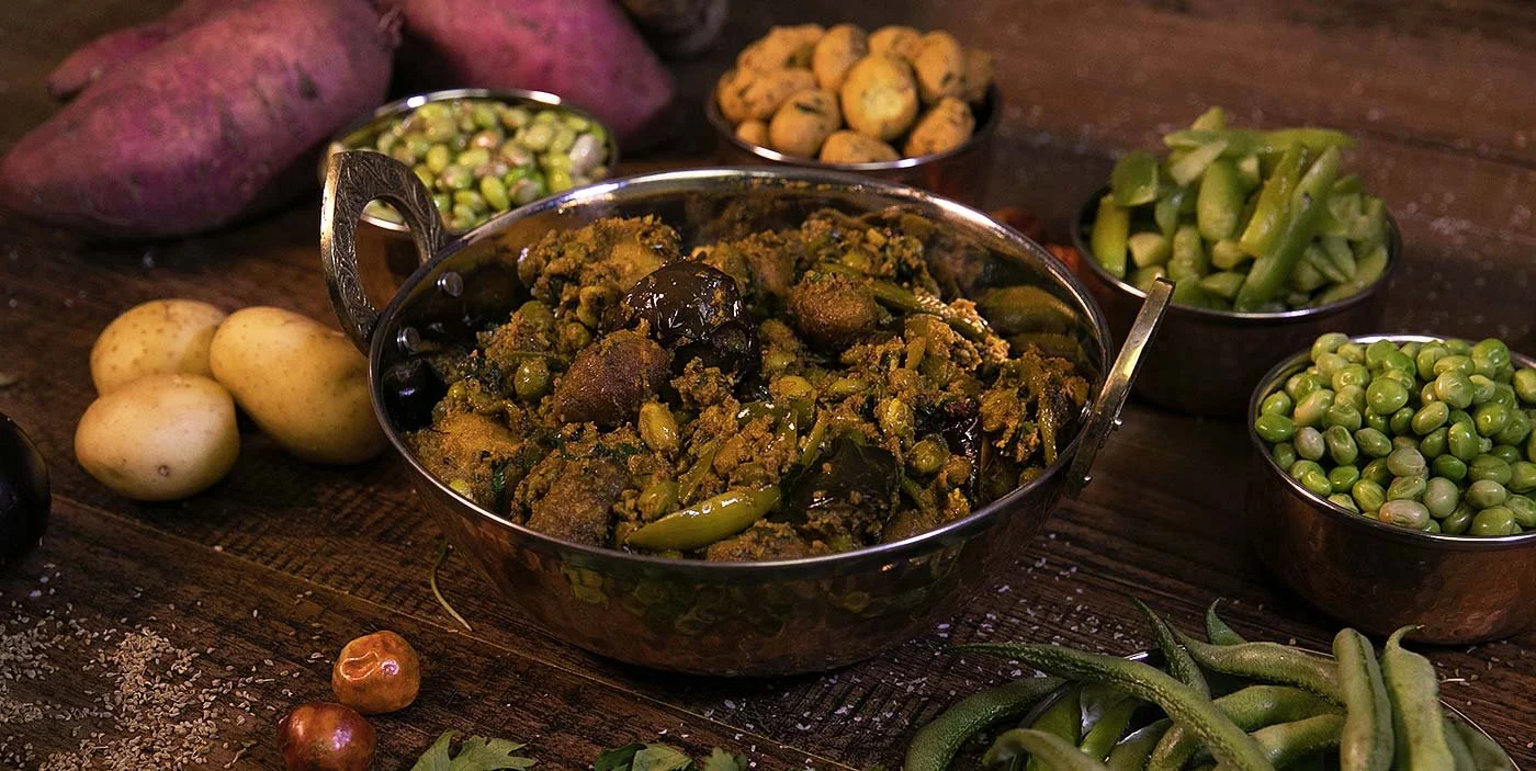 A variety of fresh vegetables on a wooden table, including sweet potatoes, potatoes, green beans, peas, and a spicy Indian vegetable dish in a stainless steel bowl.