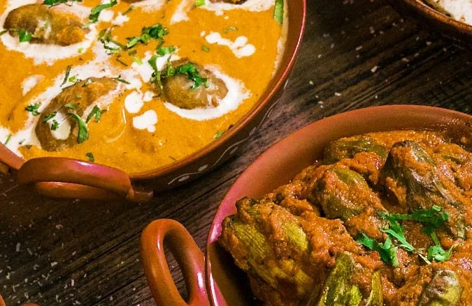 Close-up view of two Indian curry dishes served in traditional terracotta bowls, one with a creamy orange-colored curry with meatballs topped with cilantro and white sauce, and the other with a spicy green curry with vegetables.