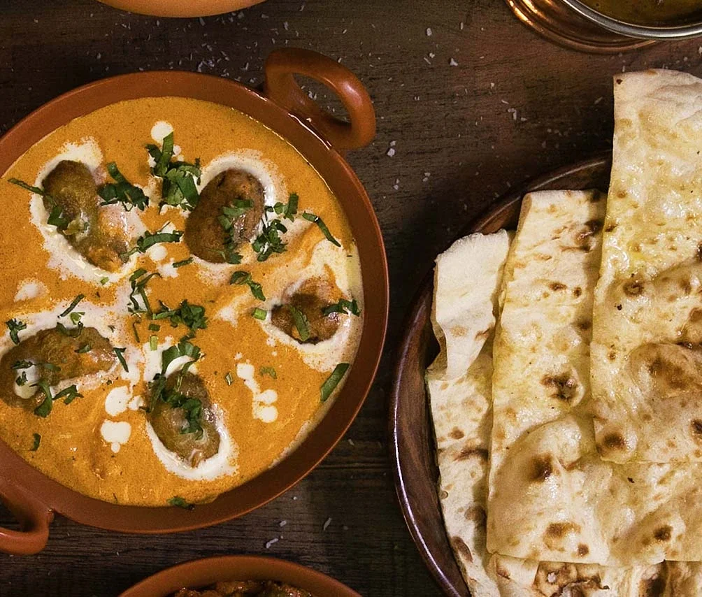 A bowl of butter chicken with grilled chicken pieces garnished with chopped cilantro, next to a plate of garlic naan bread. In the background, there is a glass of dark soda.