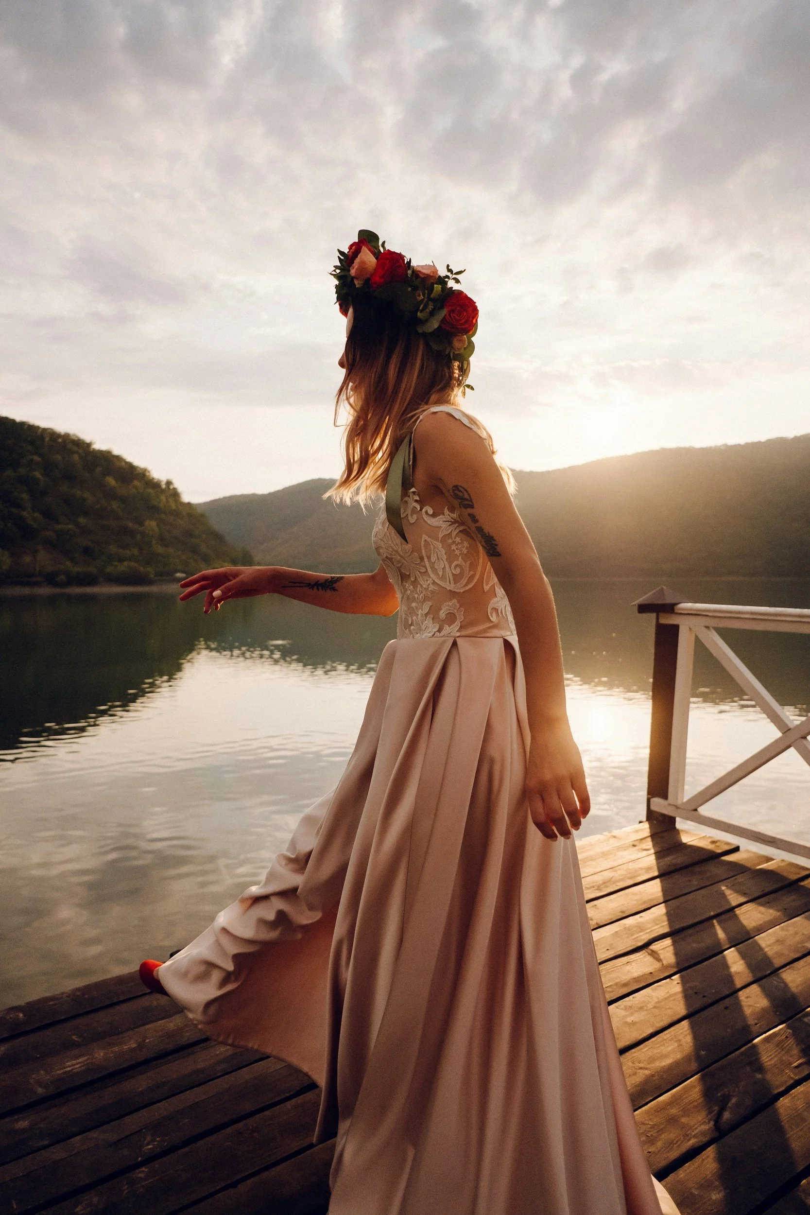 A woman wearing a floral crown and a detailed, elegant gown stands on a wooden dock by a lake at sunset, with mountains in the background.