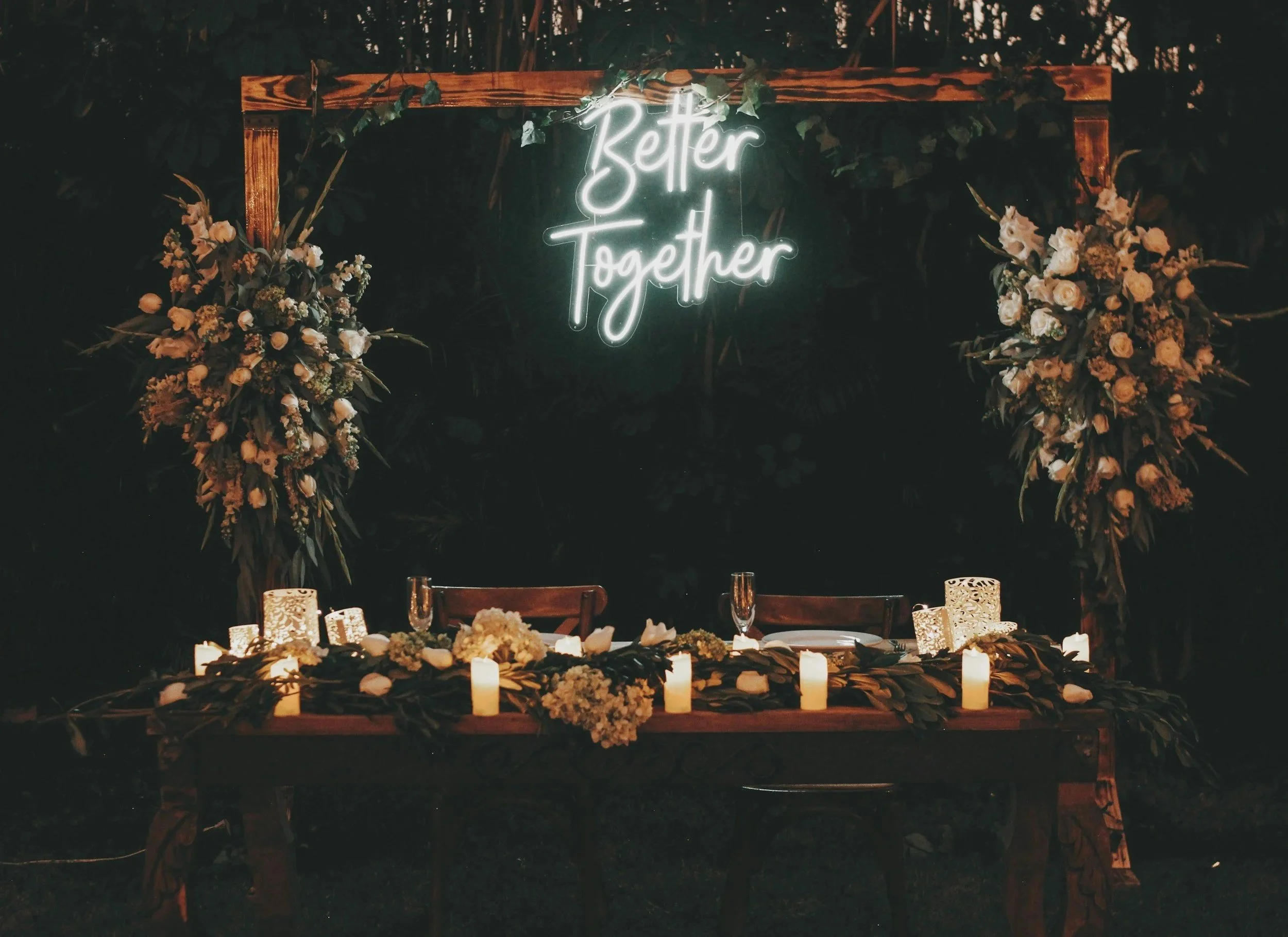 A decorated table with candles and floral arrangements, illuminated by a neon sign that says 'Better Together', set against a dark background with greenery.