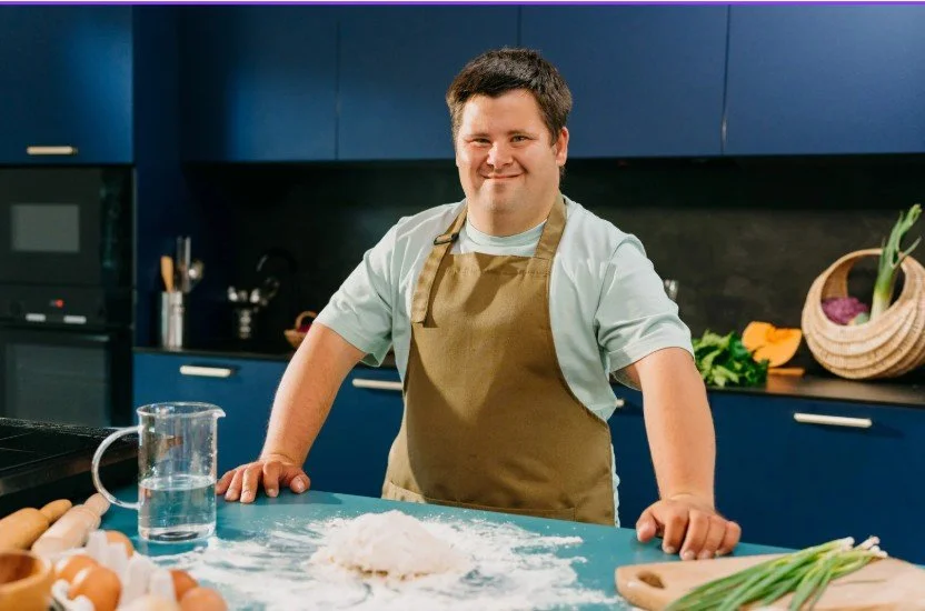 A man with Down syndrome smiling in a kitchen, wearing an apron, surrounded by baking ingredients like flour, eggs, and green onions.