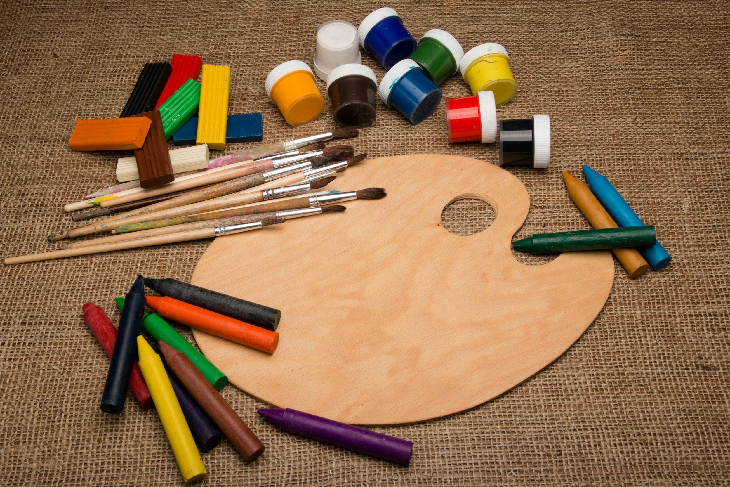 An artist's wooden palette surrounded by various art supplies including colorful chalk, paint bottles, paintbrushes, and oil pastels on a burlap surface.
