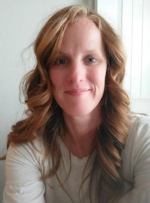 A woman with wavy red hair and fair skin smiling at the camera, wearing a white shirt, with a window in the background.