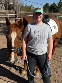 A woman standing outdoors next to a brown and white horse, holding the horse's lead rope, with a background of a fenced outdoor area and trees.