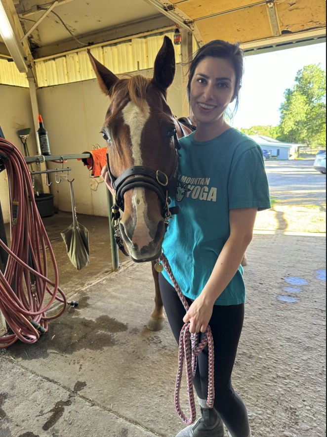 Young woman holding a horse on a lead inside a stable or barn, with outdoor scenery visible through an open door.