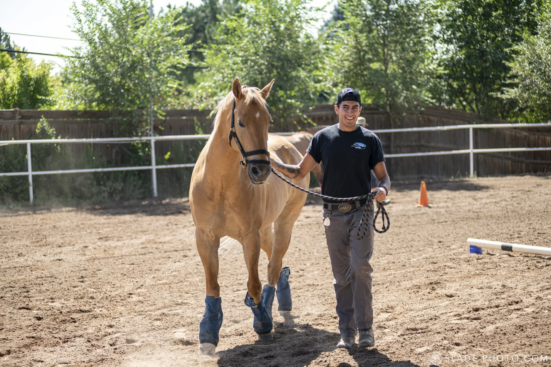 Young man guiding a tan horse with protective boots on its legs in an outdoor riding arena during a The Right Step Inc. veterans lesson. 