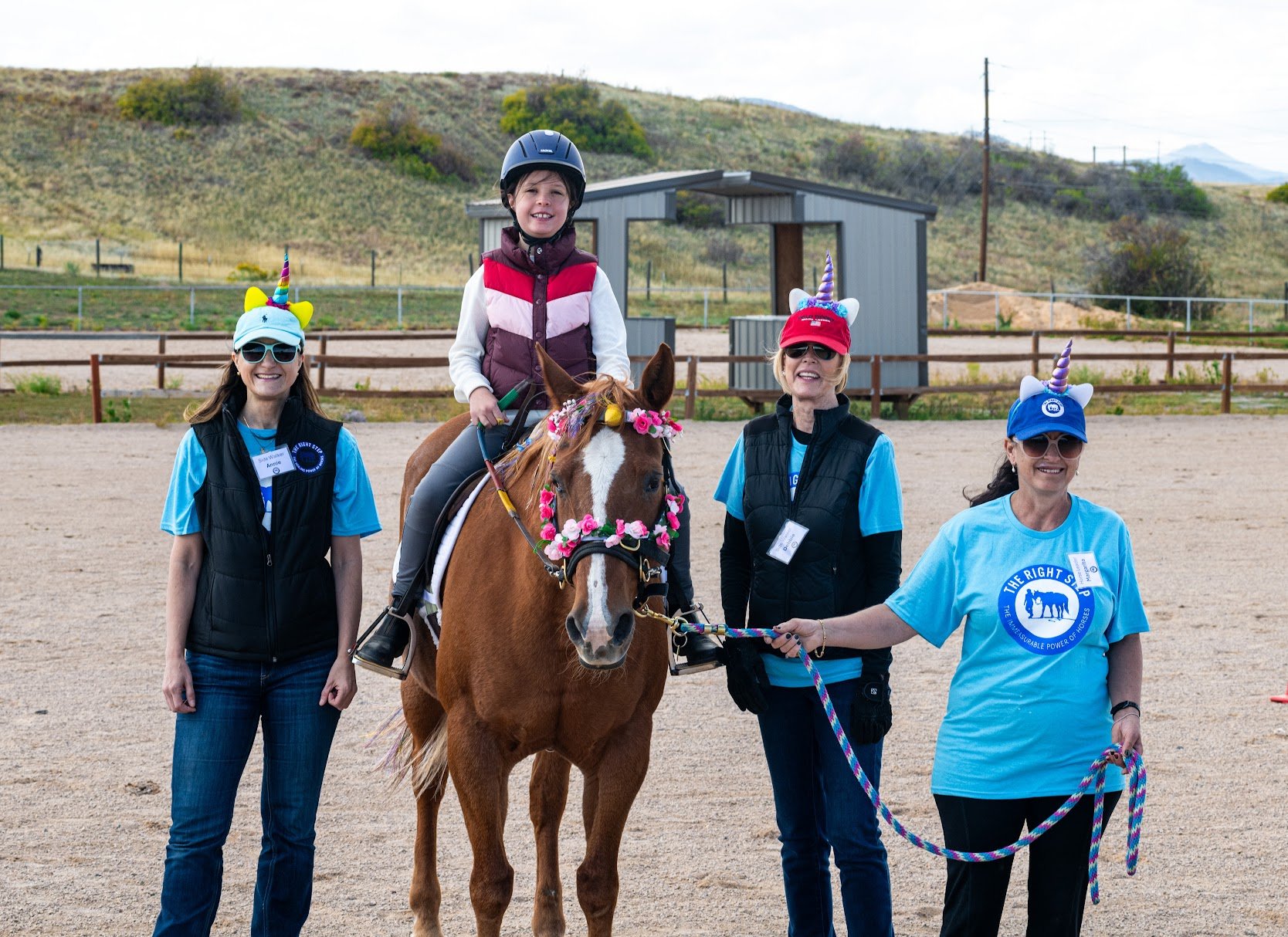 A young girl riding a TRS Unicorn Horse Show, with three volunteers standing next to her. The women are wearing unicorn headbands and sunglasses, and the girl is smiling with a helmet on.