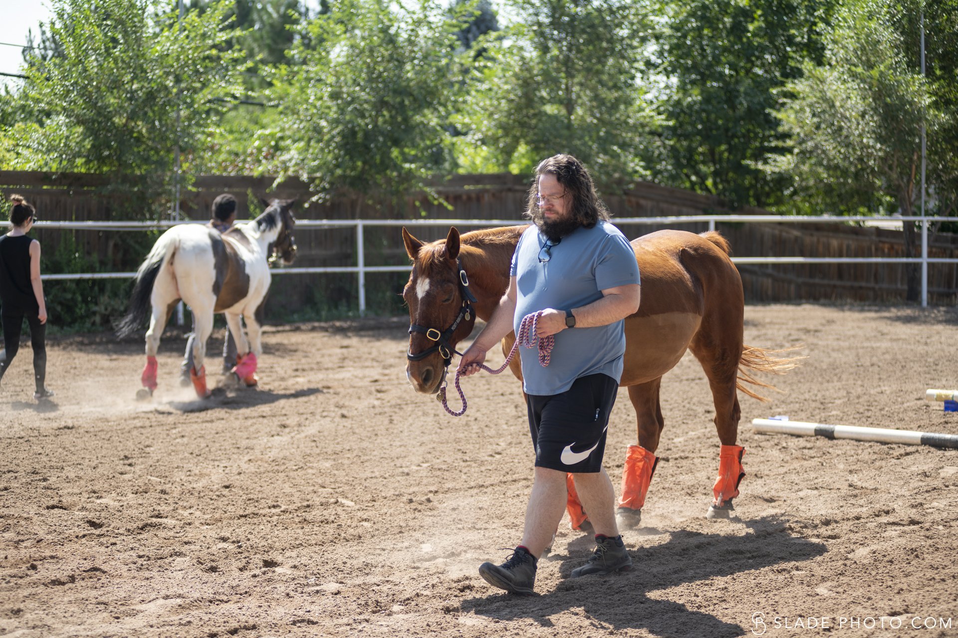 Man walking a brown horse in a sandy fenced outdoor area at The Right Step Inc. with two people and a white and black horse in the background, surrounded by green trees.