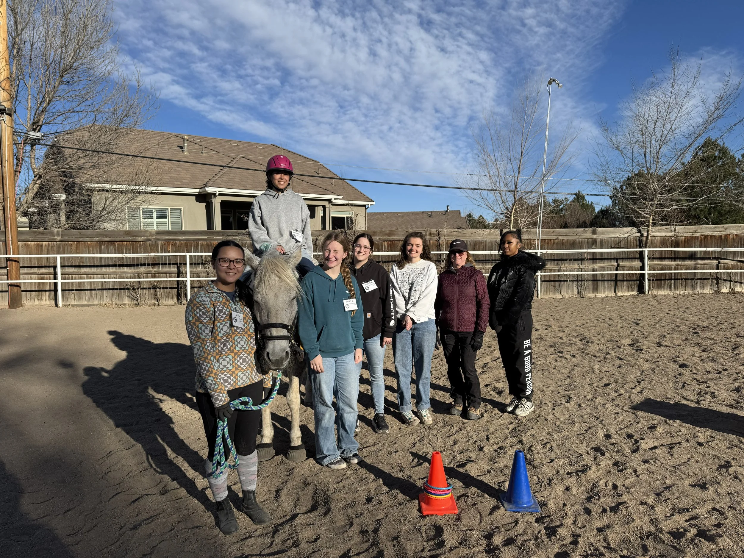 Group of six young women volunteers on a pony in an outdoor riding arena on a sunny day.