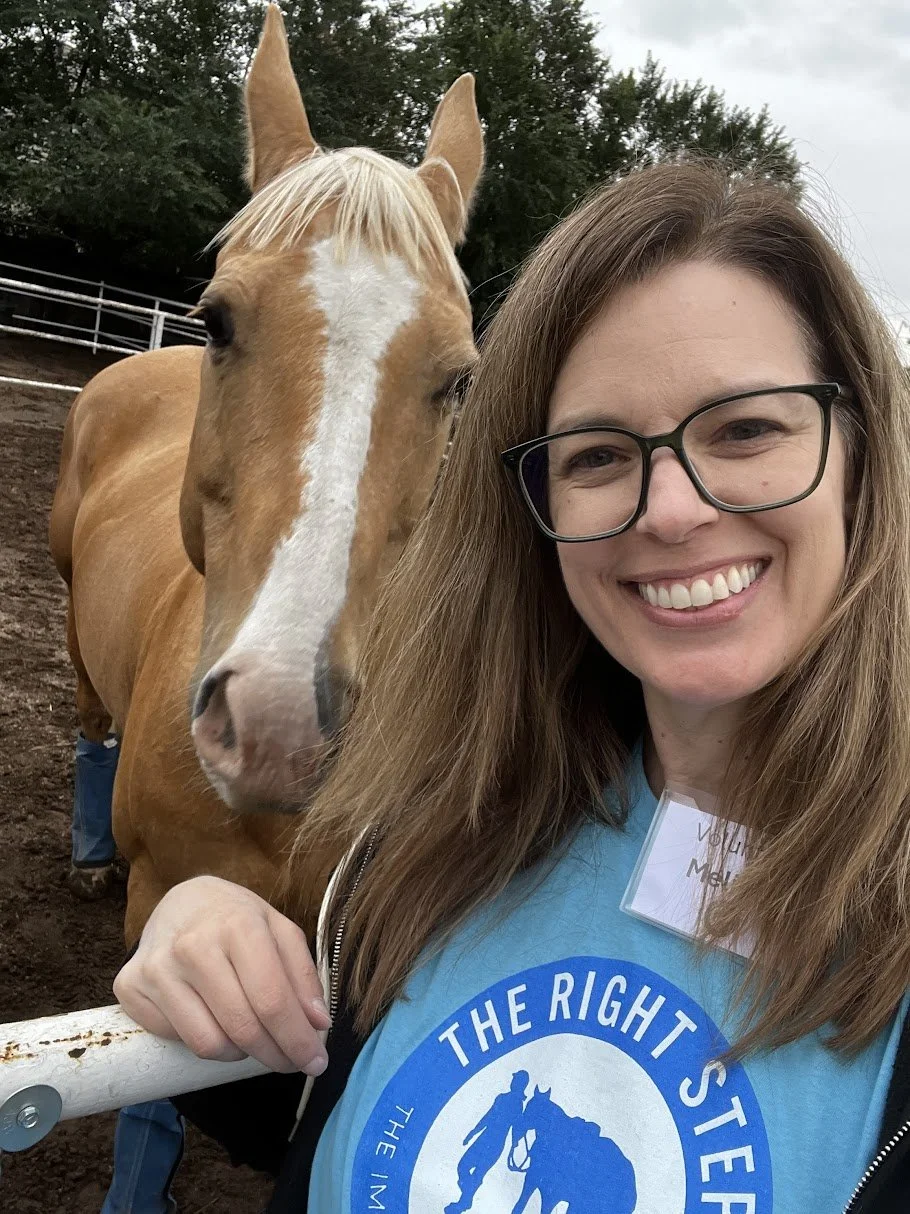 A woman smiling in front of a horse at a farm or ranch, wearing glasses and a blue shirt with a logo, on a cloudy day.