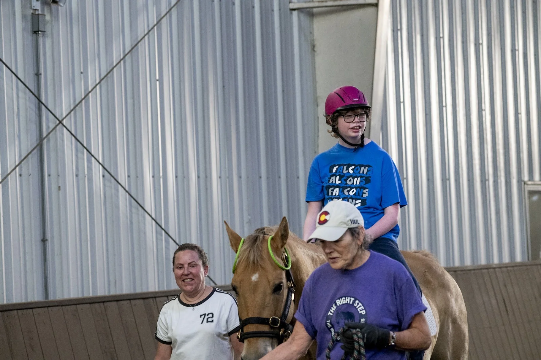 A young boy with glasses and a red helmet riding a horse guided by two women indoors. One woman is wearing a white shirt and the other a purple shirt and white cap. The background features metal walls.