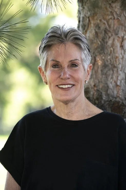 A smiling middle-aged woman with short gray hair standing outdoors near a tree, wearing a black shirt, with a blurred green background.