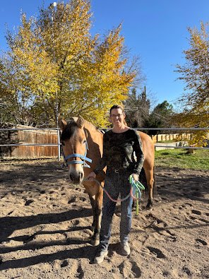 A person standing next to a brown horse with a blue halter in an outdoor riding arena, with trees displaying fall foliage in the background.
