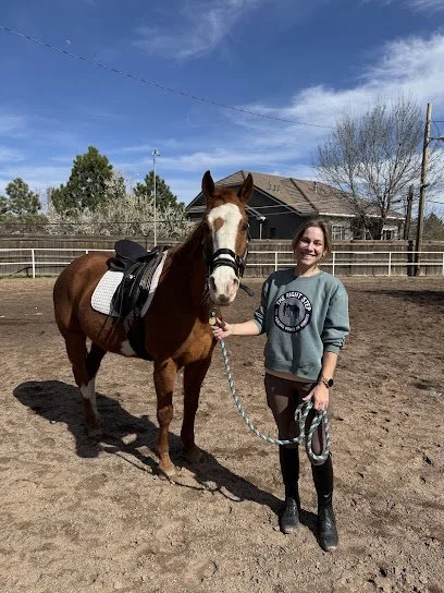 A woman standing outside in a dirt riding arena, holding a saddle horse's reins. The horse is equipped with a saddle and bridle, and the woman is smiling and wearing a sweatshirt and boots. In the background, there are trees, a house, and a clear blue sky.