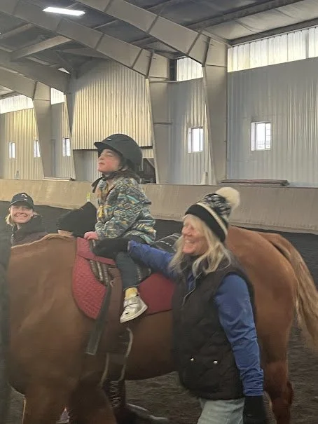 A young girl riding a horse in an indoor riding arena, with a woman walking alongside and holding her safety strap, while another girl watching and smiling.