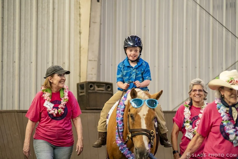 A young boy wearing a helmet rides a horse decorated with blue sunglasses and a lei, while smiling and sitting upright. Three volunteers, all wearing pink shirts, leis, and hats, stand around him smiling, inside a large indoor space with metal walls.