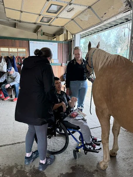 A young man in a wheelchair talking to a woman standing in front of him, with another woman and a horse nearby inside The Right Step Inc. barn.