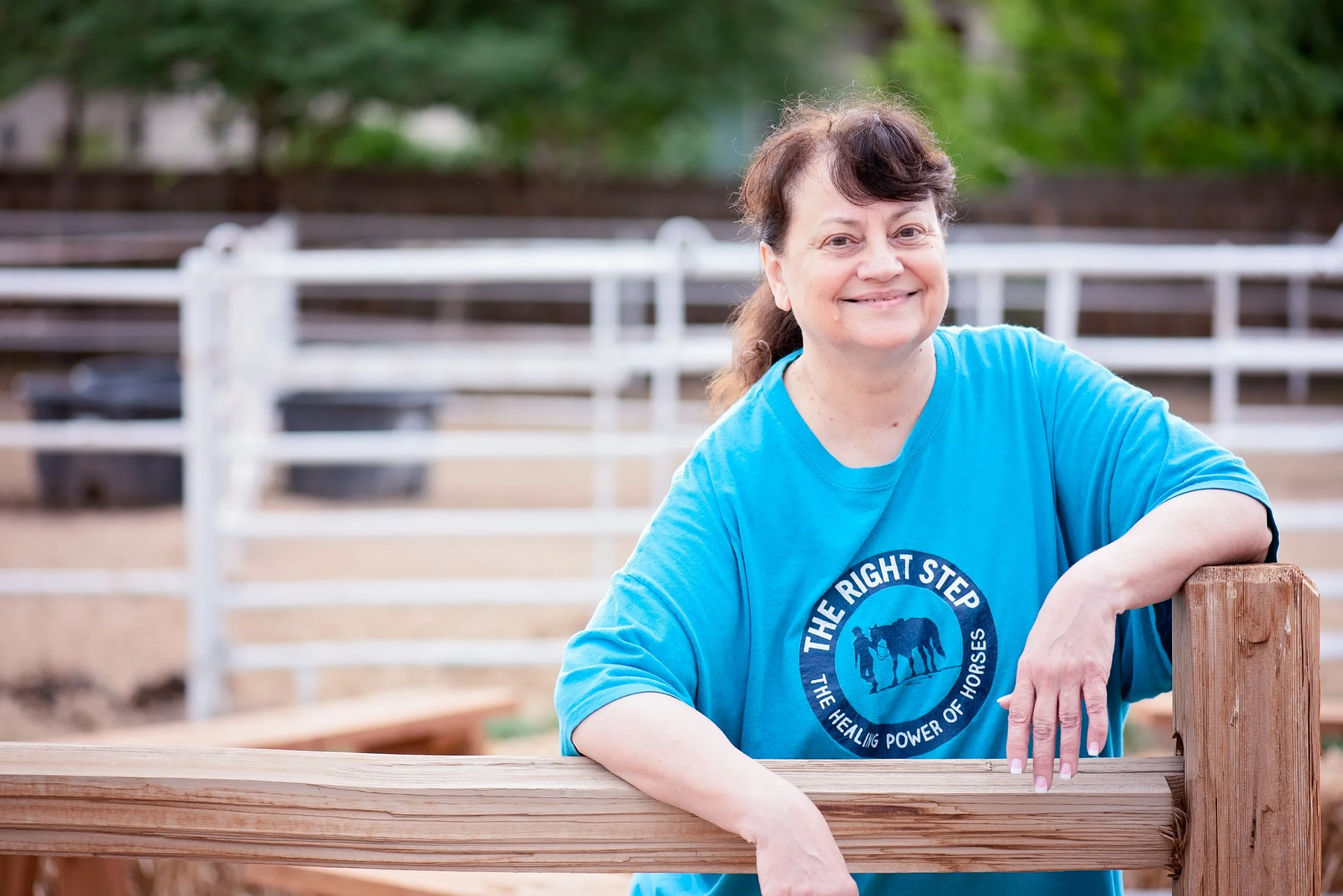Woman with curly hair smiling, wearing a blue shirt, leaning on a wooden fence at a farm or ranch.