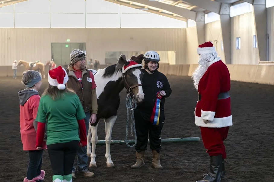 People in festive attire with a horse inside an indoor riding arena at The Right Step Inc. holiday horse show, celebrating with Santa Claus.