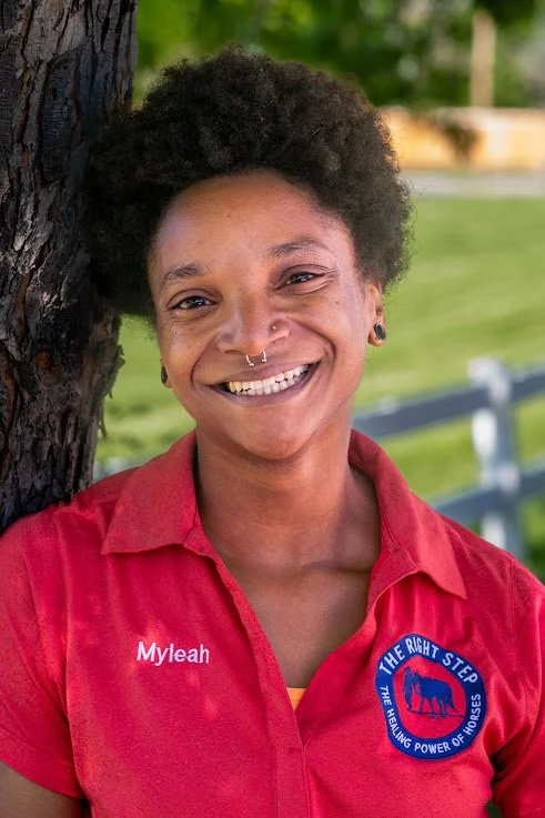 A smiling woman with short curly hair and a nose piercing, wearing a red shirt with 'Myleah' embroidered on it and a patch that reads 'The Right Step' with an elephant logo, standing outdoors beside a tree.