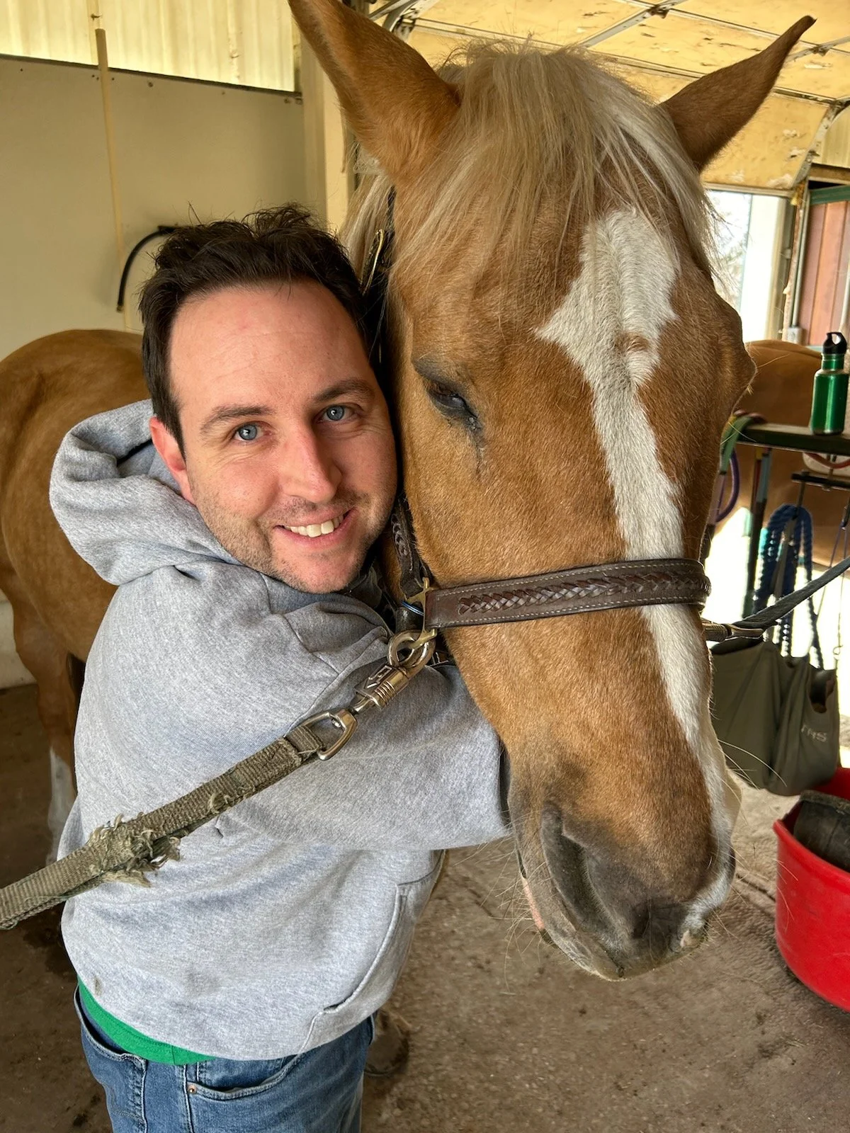 A smiling man hugging a light brown horse with a white blaze on its face inside The Right Step Inc. barn. 