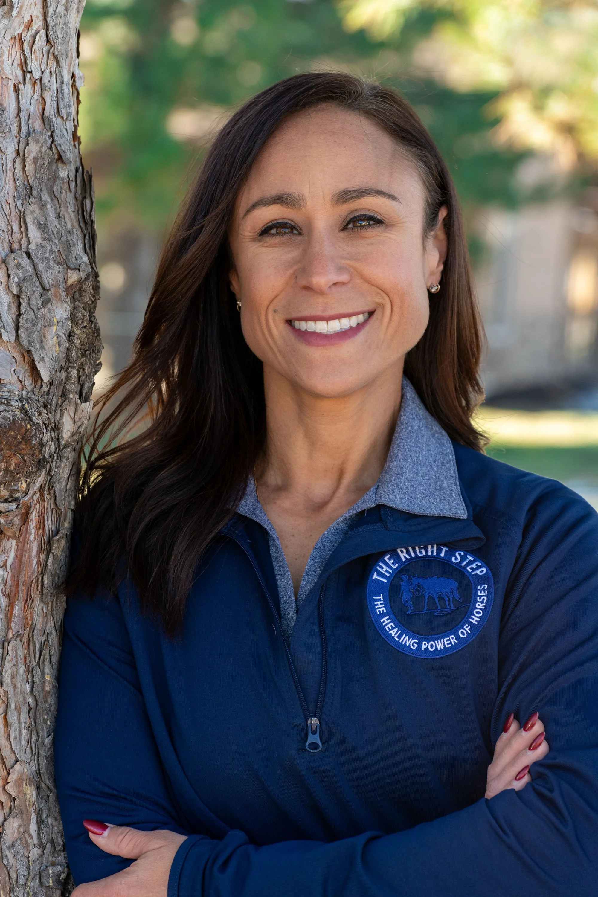 A woman with long dark hair smiling outdoors, wearing a blue jacket with a logo and cross arms, standing next to a tree.