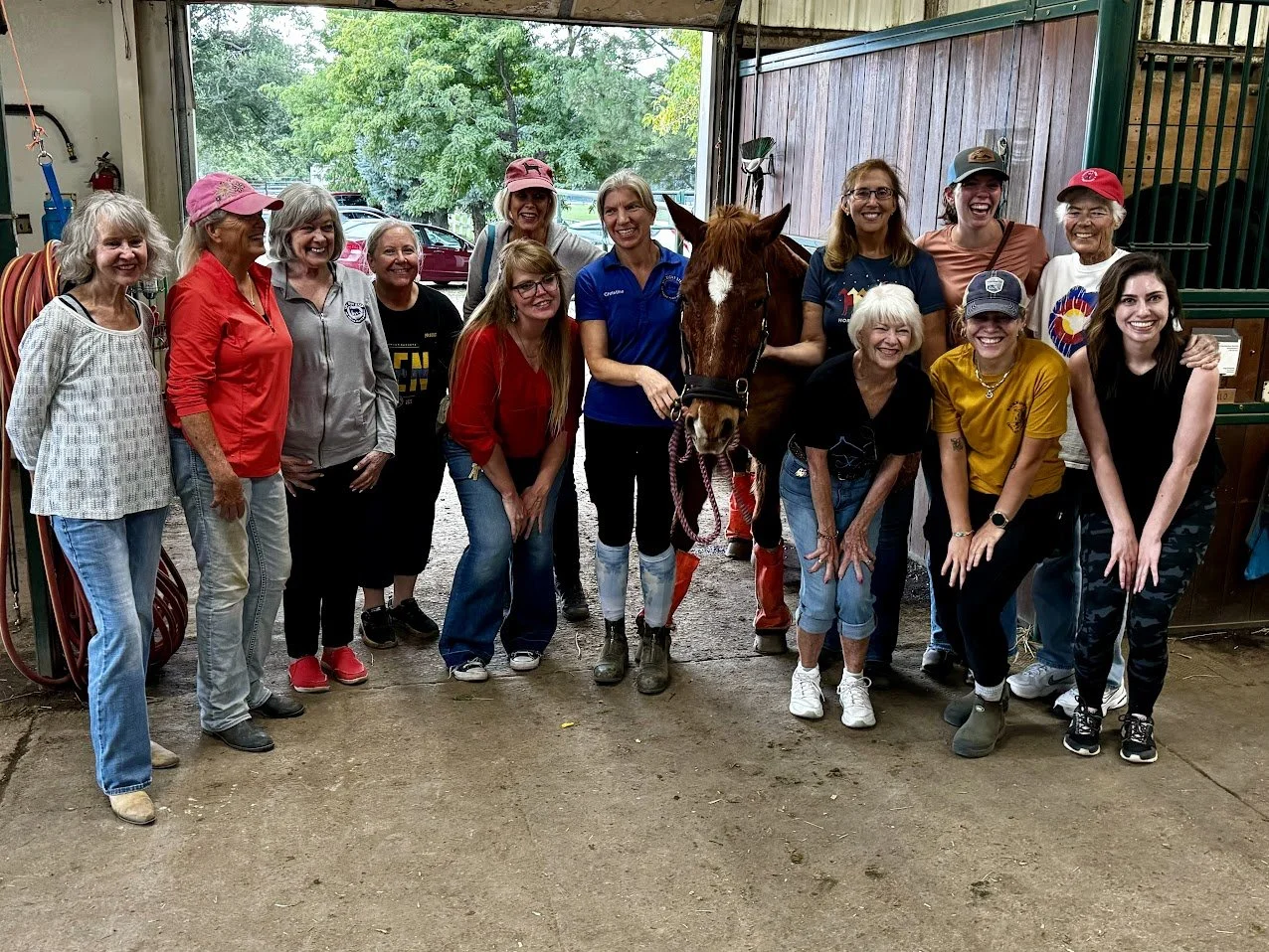 A group of women and girls gathered inside a horse stable, with a horse at the center. They are smiling and at The Right Step Inc. barn. 