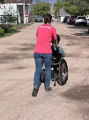 A woman wearing a pink volunteer shirt pushing a person in a wheelchair down a gravel road.